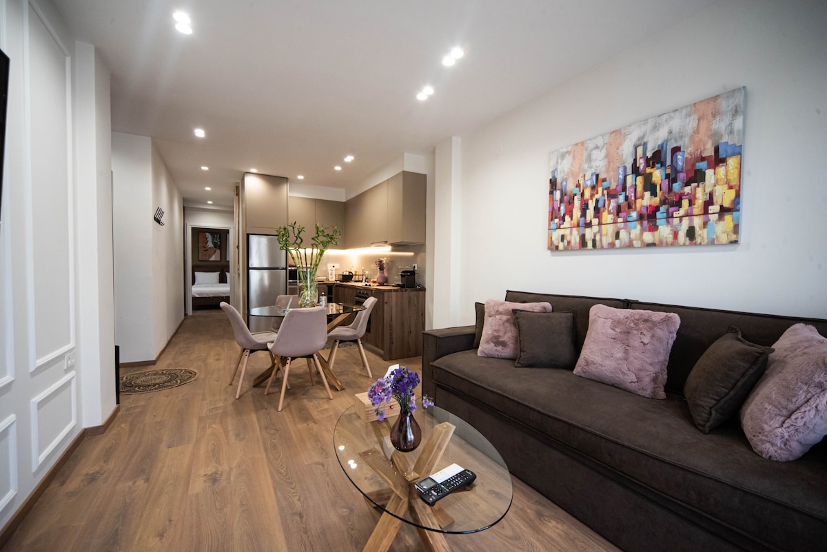 A modern living area features a dark sofa adorned with pink throw pillows, complemented by a glass coffee table. Two chairs are arranged around the table, and a colorful wall art piece adds a touch of vibrancy. Soft lighting highlights the open layout leading to the kitchen.