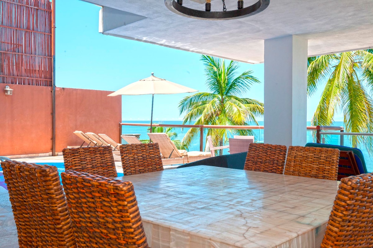 An outdoor dining area is visible, featuring a large, tiled table surrounded by wicker chairs. The scene includes a private pool in the background, with comfortable lounge chairs and a white umbrella under a clear blue sky, framed by palm trees and ocean views.