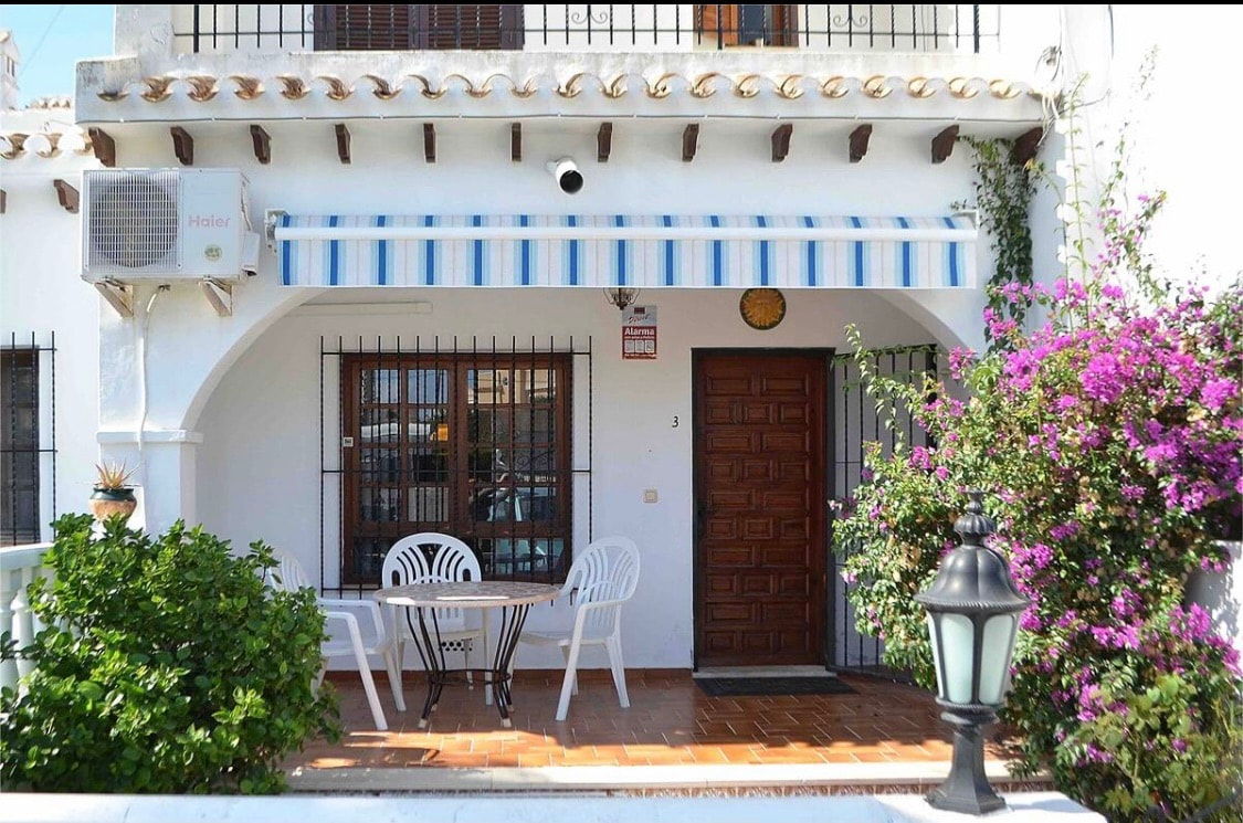 A charming entrance features a wooden door framed by lush greenery and flowering plants. A small table and two chairs are positioned under a striped awning, providing a shaded area for relaxation. An outdoor lamp adds illumination to the inviting space.