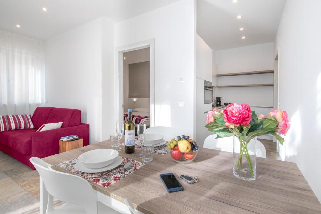 A modern dining area is featured, with a wooden table set for four, decorative tablecloth, and a centerpiece of vibrant flowers and fresh fruit. A cozy red sofa is visible in the background, with natural light filtering through sheer curtains.