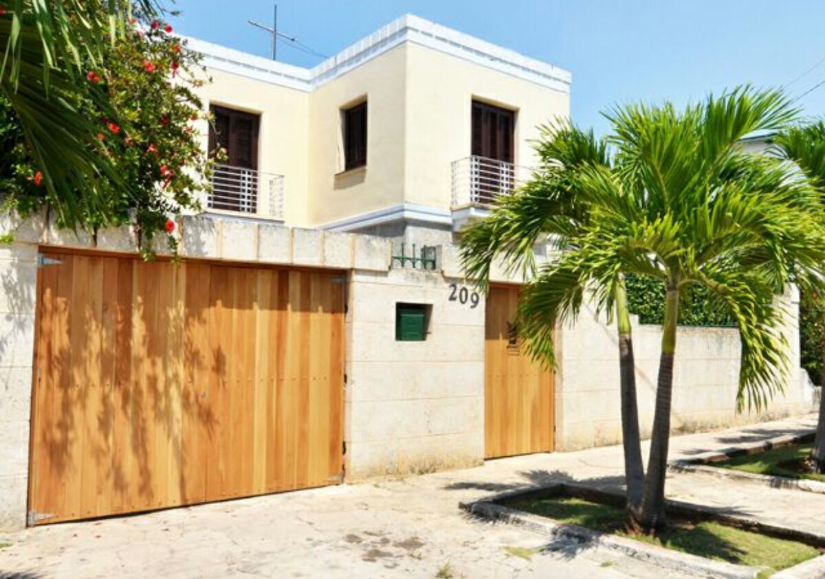 The exterior of the property features a tall stone wall with wooden double gates, providing a warm entrance. The house is adorned with light-colored walls and has a two-story design. Lush palm trees and vibrant foliage are visible, enhancing the greenery of the surroundings.