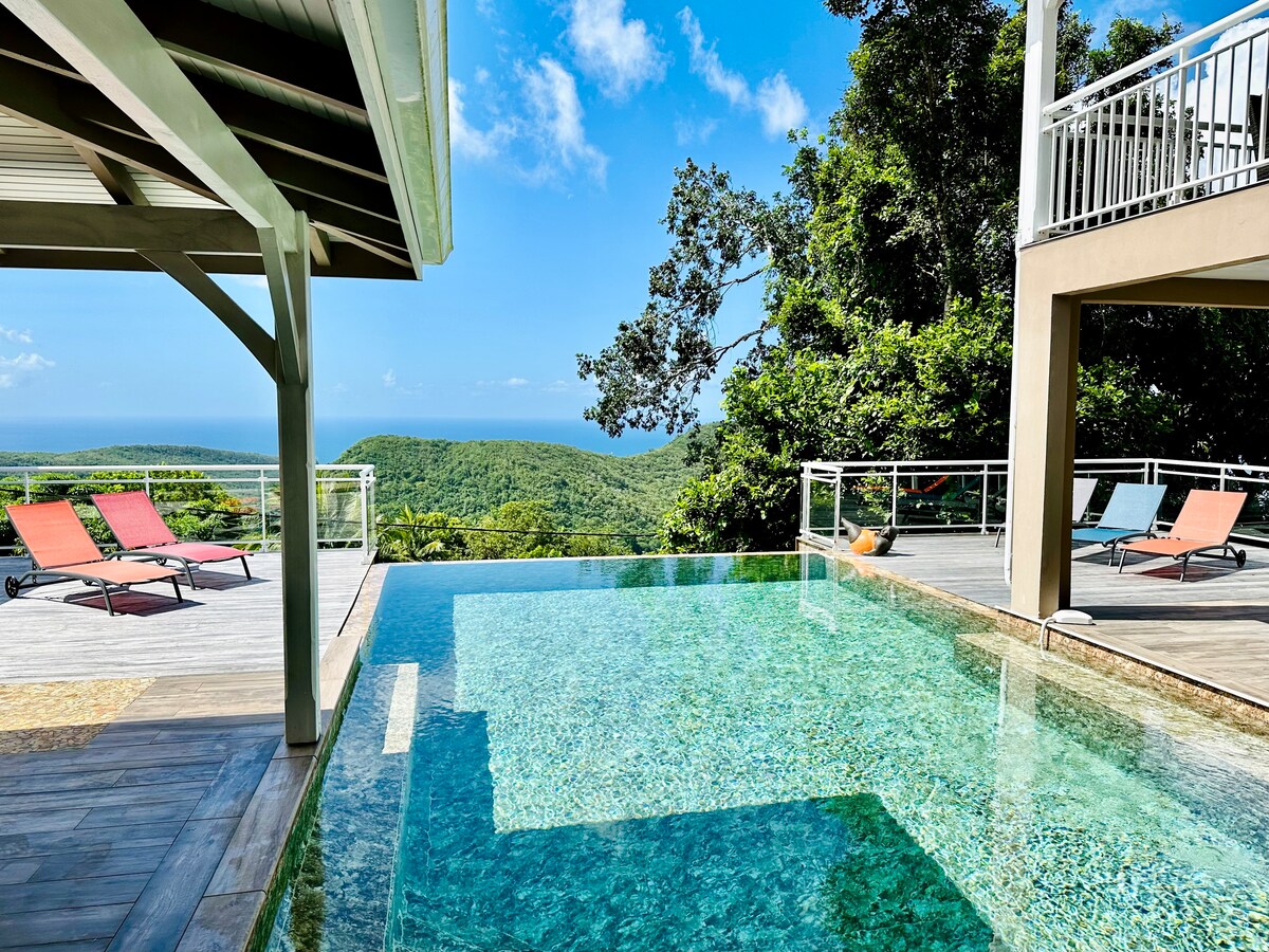 An infinity pool reflects the clear blue sky, surrounded by a spacious wooden deck with colorful loungers. Lush greenery and distant hills are visible in the background, complementing the serene outdoor setting.