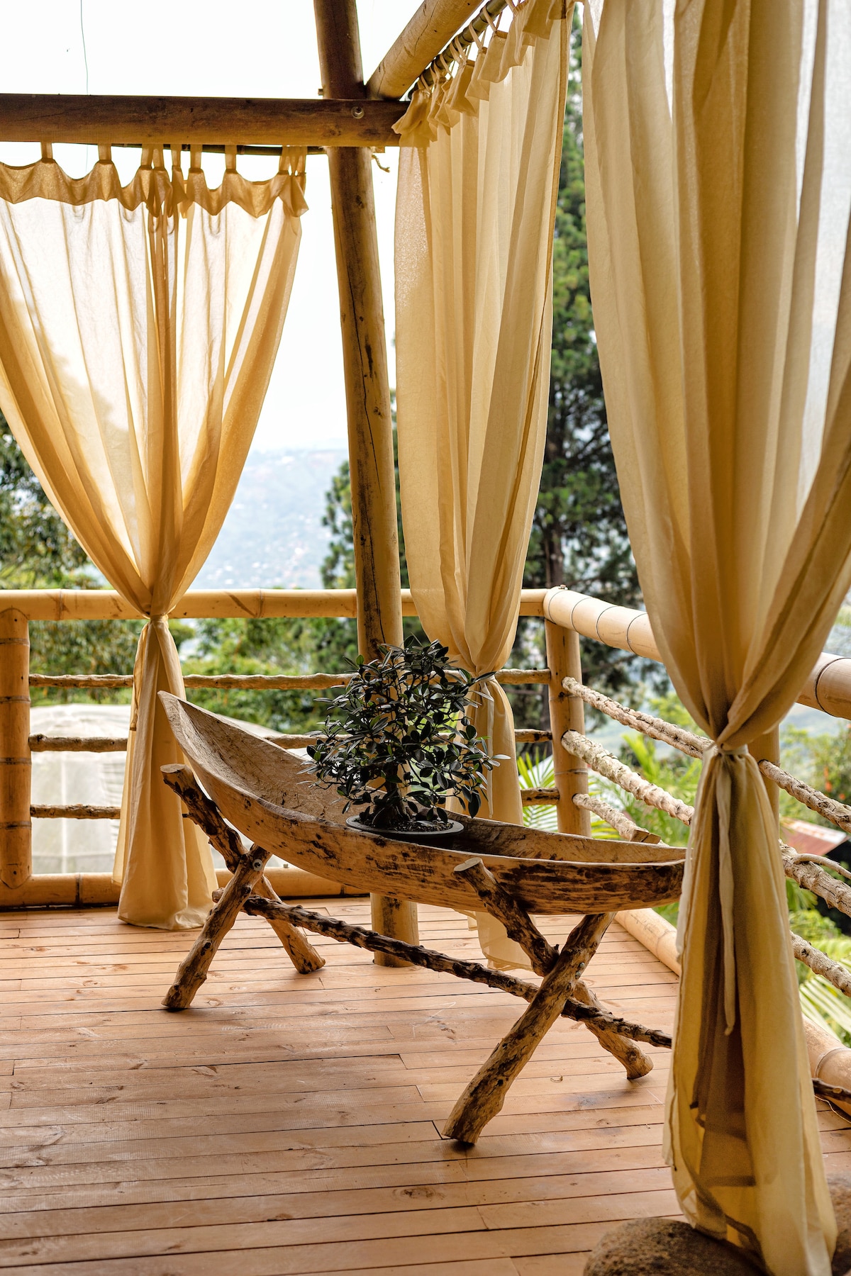 An inviting balcony is adorned with sheer beige curtains, allowing natural light to filter through. A rustic wooden table, crafted from natural materials, is positioned in the center, with a small potted plant adding a touch of greenery.