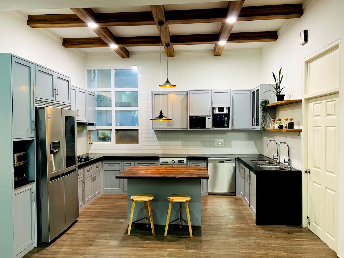 A modern kitchen features grey cabinetry with a blend of open and closed storage. Stainless steel appliances, including a refrigerator and dishwasher, are visible. A wooden island with two stools offers space for meal preparation, and natural light streams in through a large window.