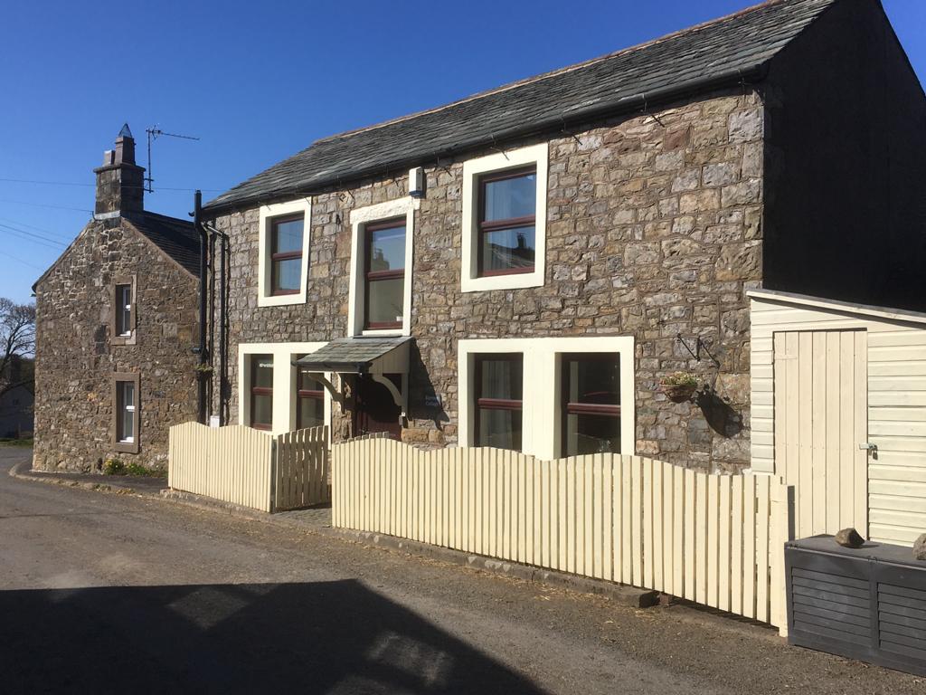 A traditional 18th-century stone cottage is featured, showcasing a wooden front fence and a welcoming entrance. Notable elements include large windows allowing natural light and a charming roof structure. The exterior reflects the character of the village, enhancing the historic ambiance of the property.