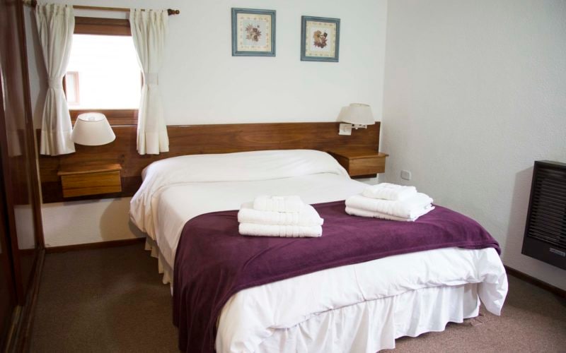A sleeping area features a spacious bed draped with a white coverlet and a dark purple blanket. Two neatly rolled towels are placed on the bed. Soft white curtains frame a window, and framed artwork is displayed on the wall above the headboard.