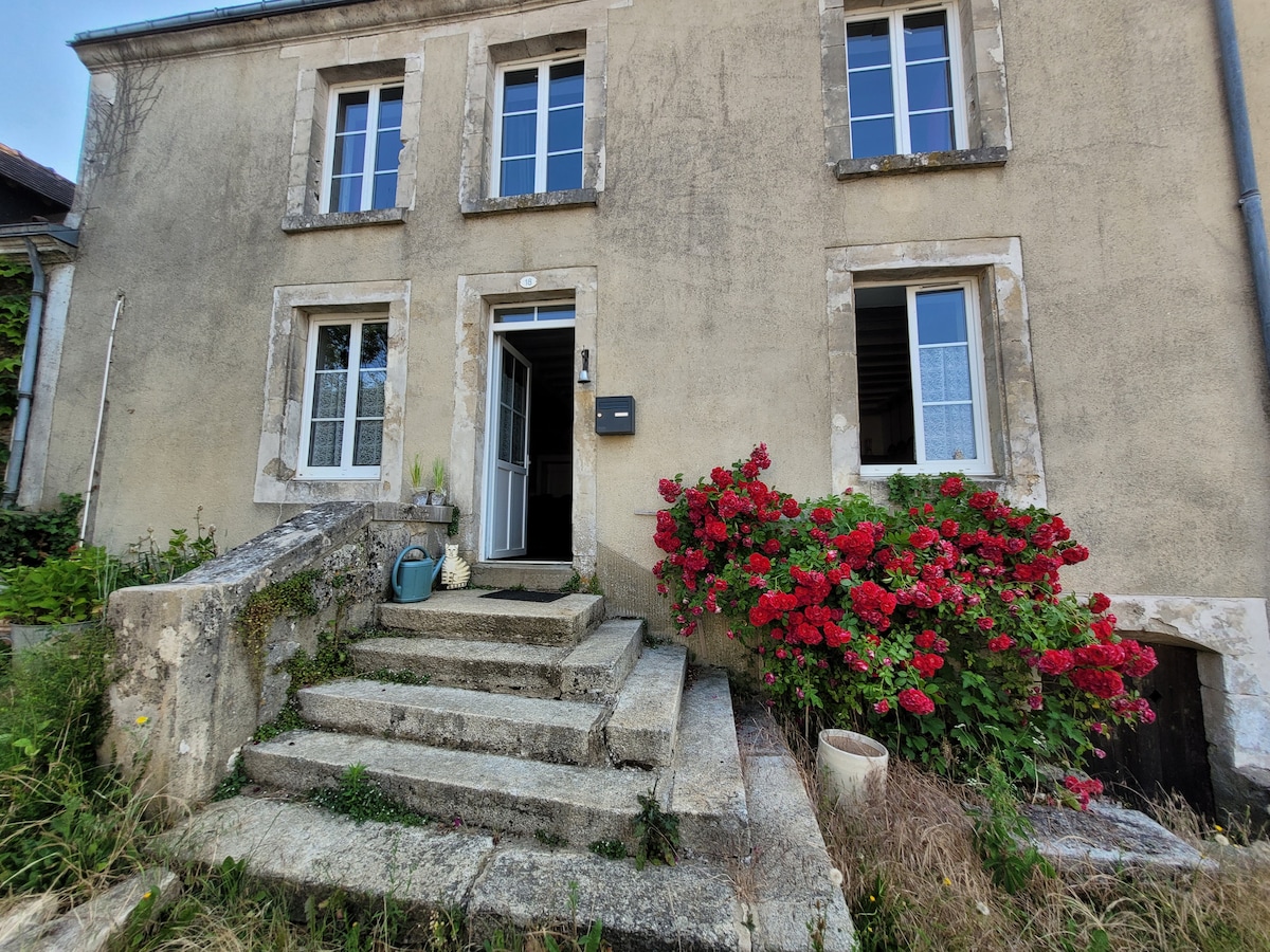 The exterior of a historic house is shown, featuring a stone staircase leading to an entrance, bordered by blooming hydrangeas. Large windows are visible, allowing natural light to enter the building, which exhibits signs of age on its façade.