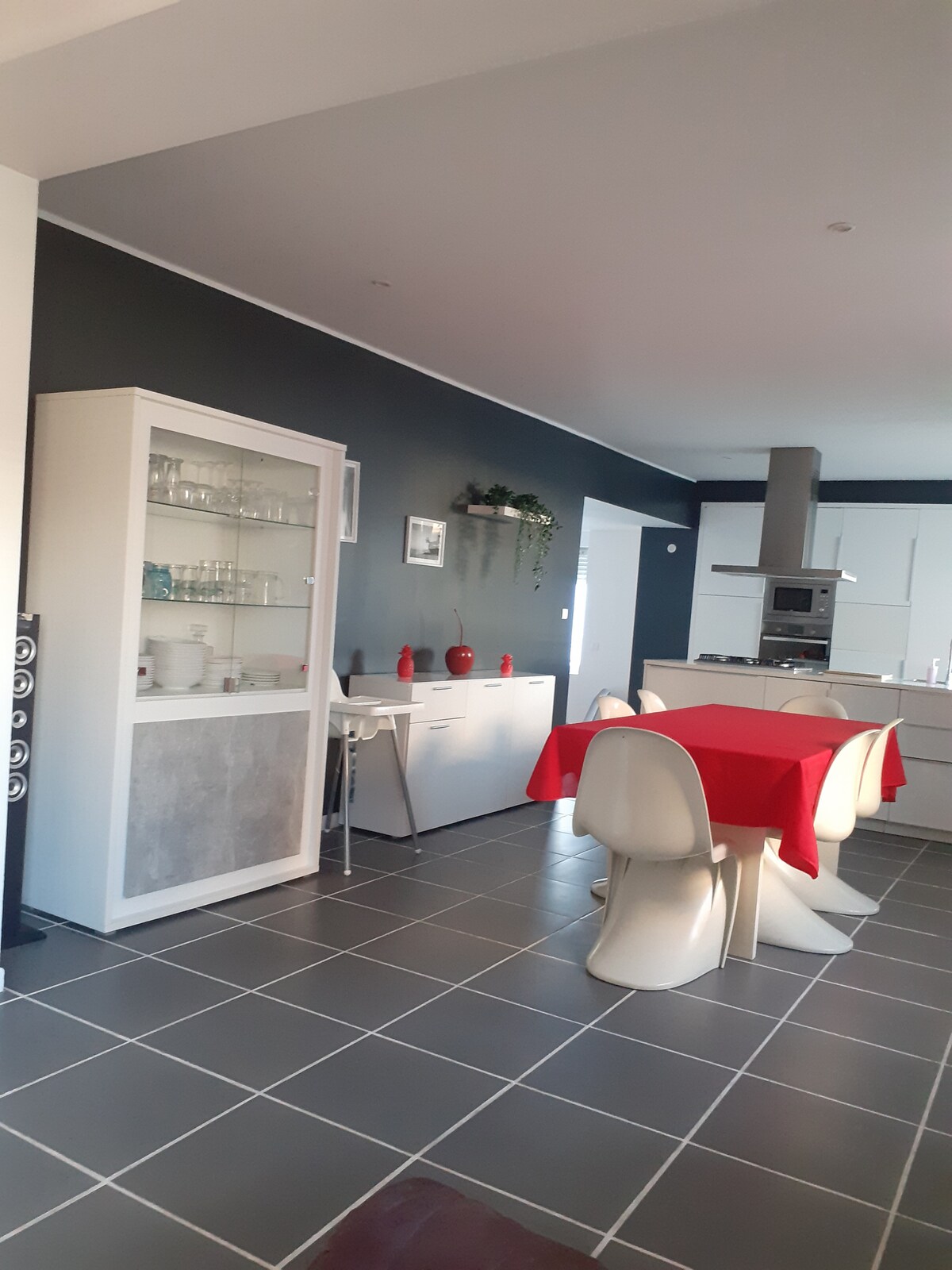 A modern kitchen space features a dining table covered with a red tablecloth, surrounded by white chairs. A glass-front cabinet displays dishware, while a sleek kitchen unit with appliances is visible in the background. Gray tiled flooring complements the charcoal walls.