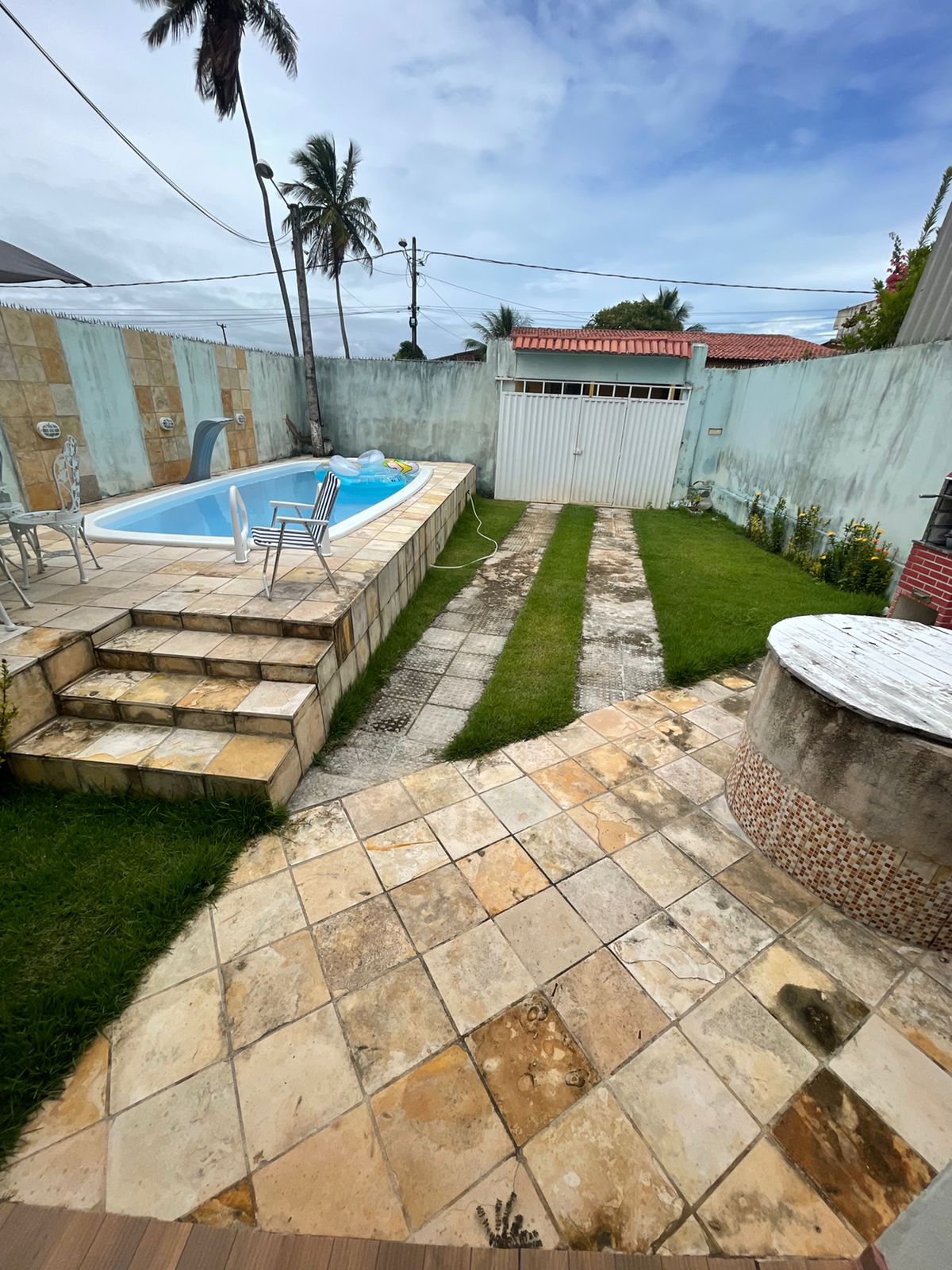 The outdoor area features a swimming pool set within stone steps, alongside a well-maintained lawn bordered by paving stones. A barbecue area is visible, with a circular stone structure. Palm trees can be seen in the background, enhancing the tropical ambiance of the space.