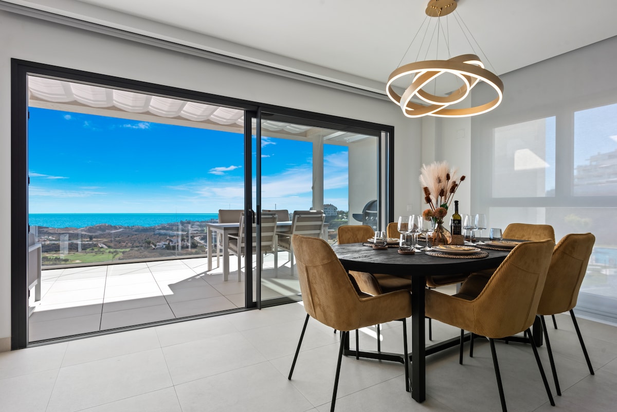 A dining area features a round wooden table, surrounded by beige upholstered chairs. Large glass doors open to reveal a panoramic view of the coastline and the sea. Natural light floods the space, enhancing the clean, modern aesthetic.