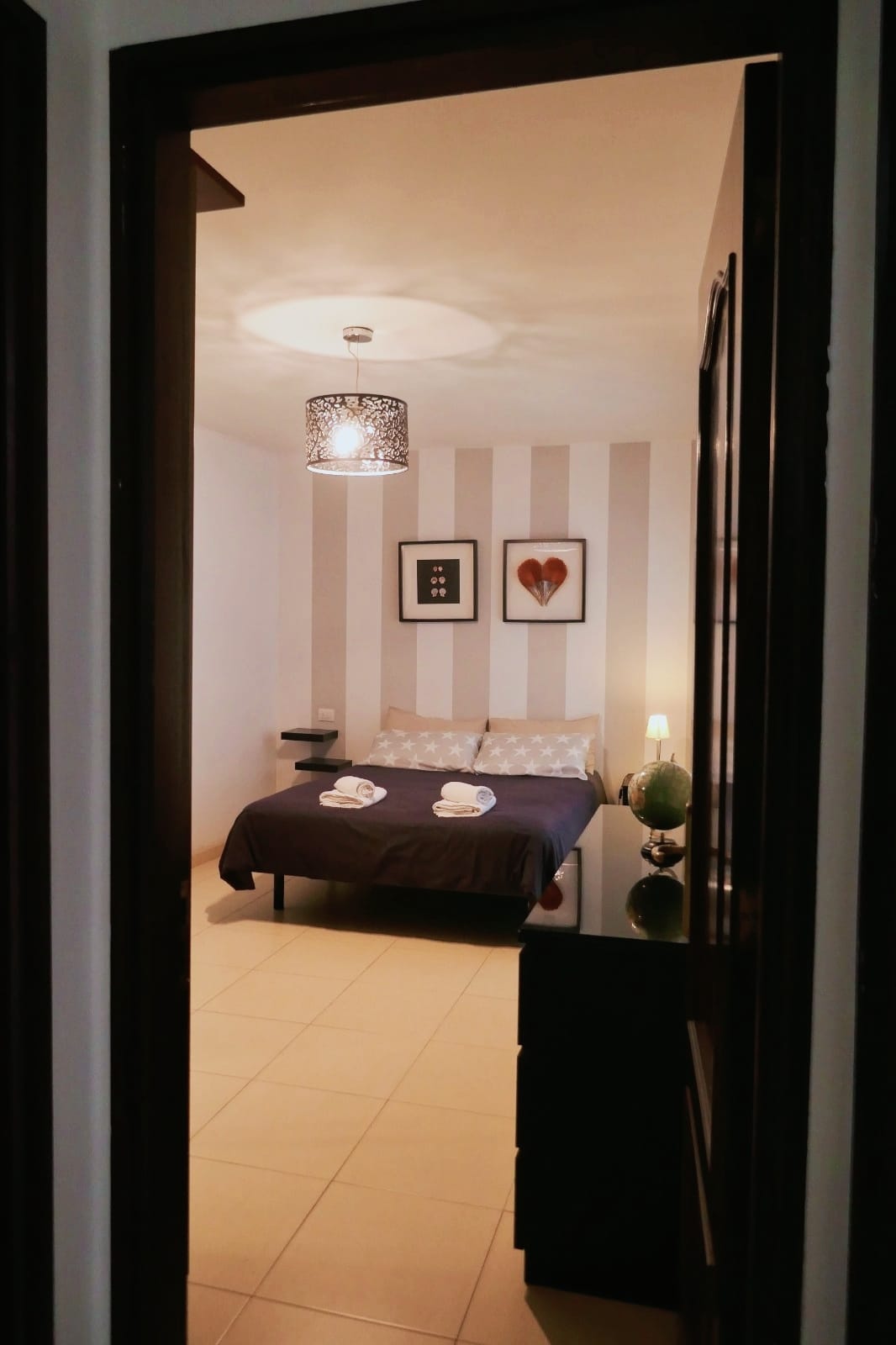 A well-appointed bedroom is visible through a door frame. Soft lighting is provided by a stylish ceiling fixture. The space features a comfortable bed adorned with coordinated linens, framed artwork on the walls, and a decorative lamp on a nightstand beside the bed.