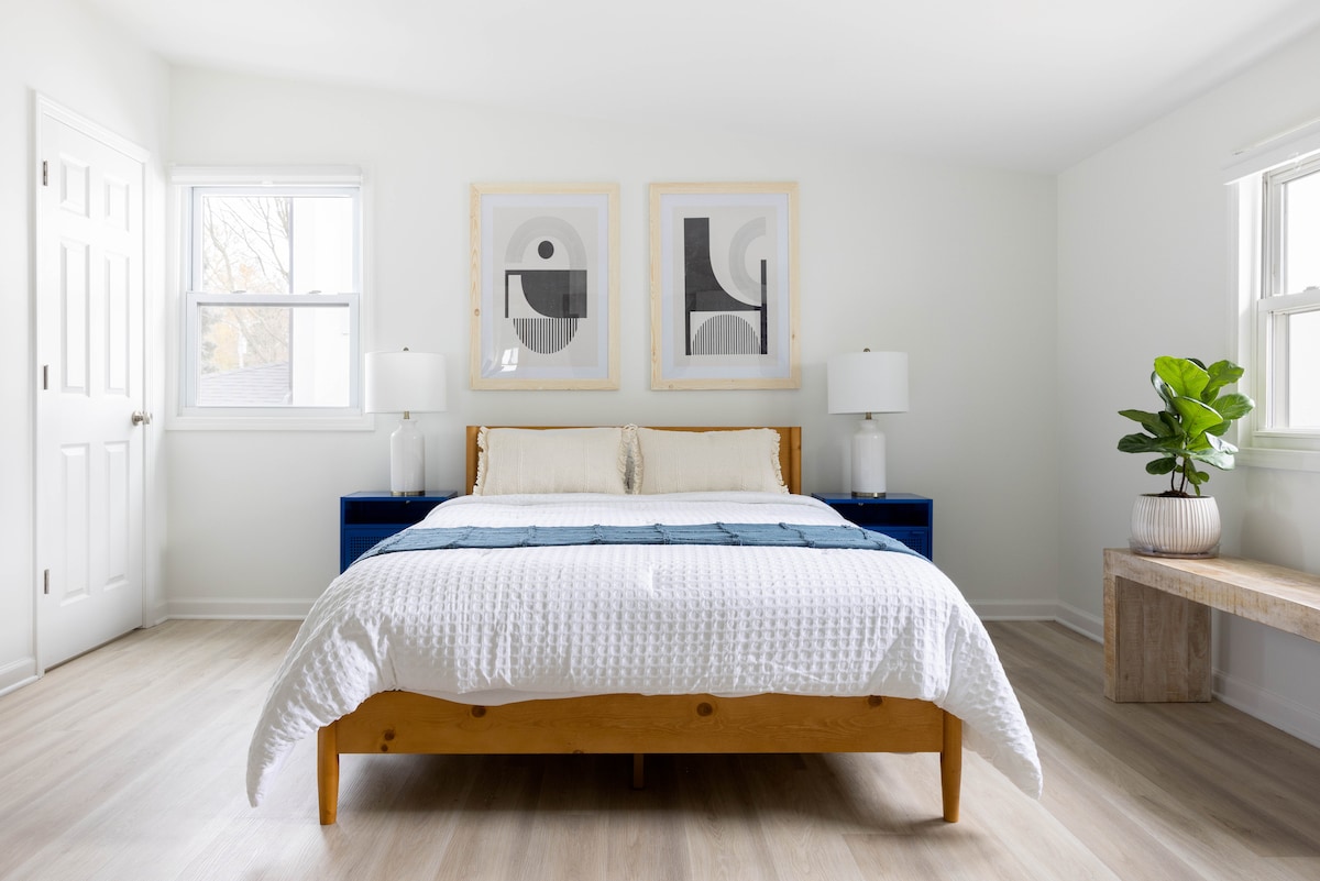 A bright bedroom features a wooden bed with a textured white duvet, complemented by two matching bedside lamps. Framed artwork adorns the walls, and a small green plant adds a touch of nature. Windows provide natural light, creating a serene atmosphere.