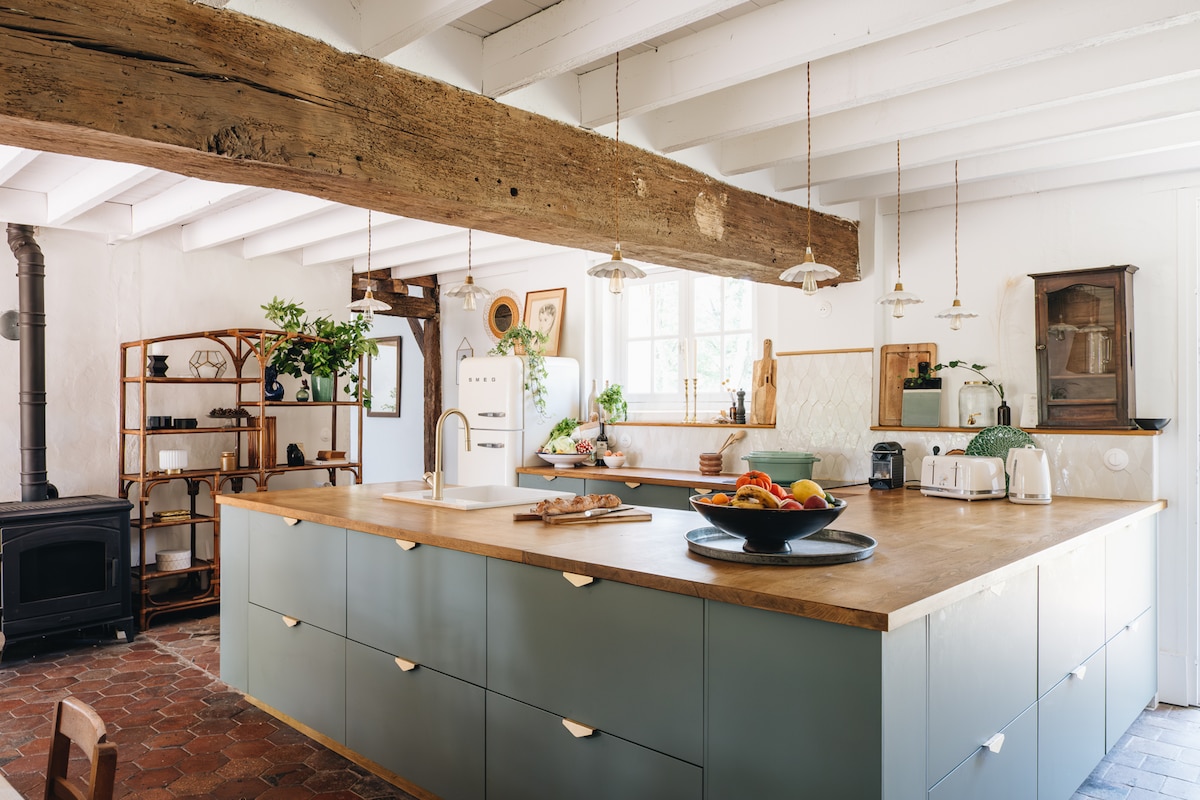 An open and inviting kitchen space features a large central island with a warm wooden countertop. Pendant lights hang above, illuminating the area, while cabinets in soft blue provide storage. A variety of plants are displayed, contributing to a natural ambiance. Essential appliances are visible.