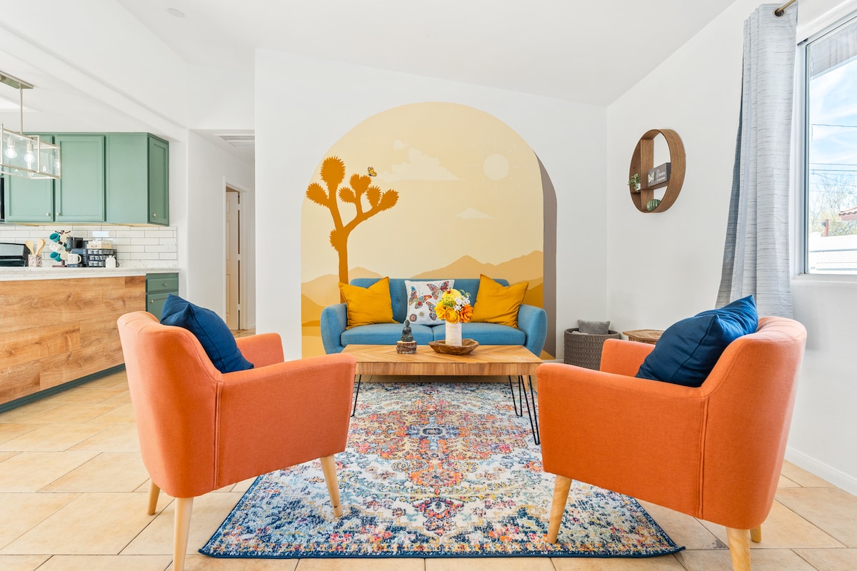 A cozy living area is presented with a vibrant blue sofa and two orange chairs, centered around a wooden coffee table adorned with a floral arrangement. A decorative mural depicting a desert scene adds character, while natural light fills the space through a window.