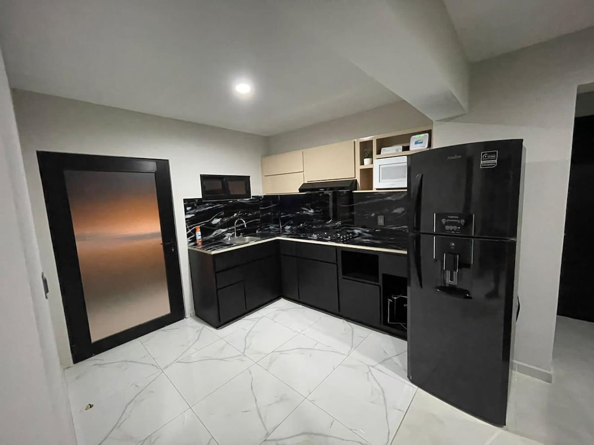 A modern kitchen is presented, featuring sleek black cabinetry and a marble countertop. Stainless steel appliances, including a refrigerator and stove, complement the dark aesthetic. Ample storage cabinets and a microwave are visible, along with a well-lit atmosphere contributed by overhead lighting.
