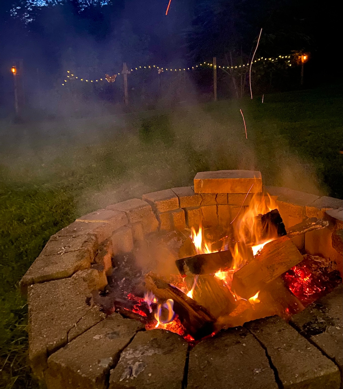 A circular stone firepit is centered in the image, with glowing logs burning brightly. Warm orange flames and glowing embers contrast with the soft green grass surrounding the pit. Twinkling lights are faintly visible in the background, creating a serene evening ambiance.