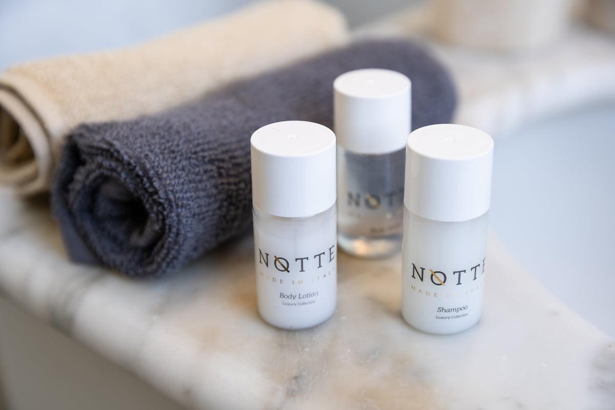 A set of travel-sized toiletries is arranged on a marble surface, including a body lotion, shampoo, and a clear liquid. Two neatly folded towels, one beige and one navy blue, are positioned beside the products, providing a simple, organized appearance.