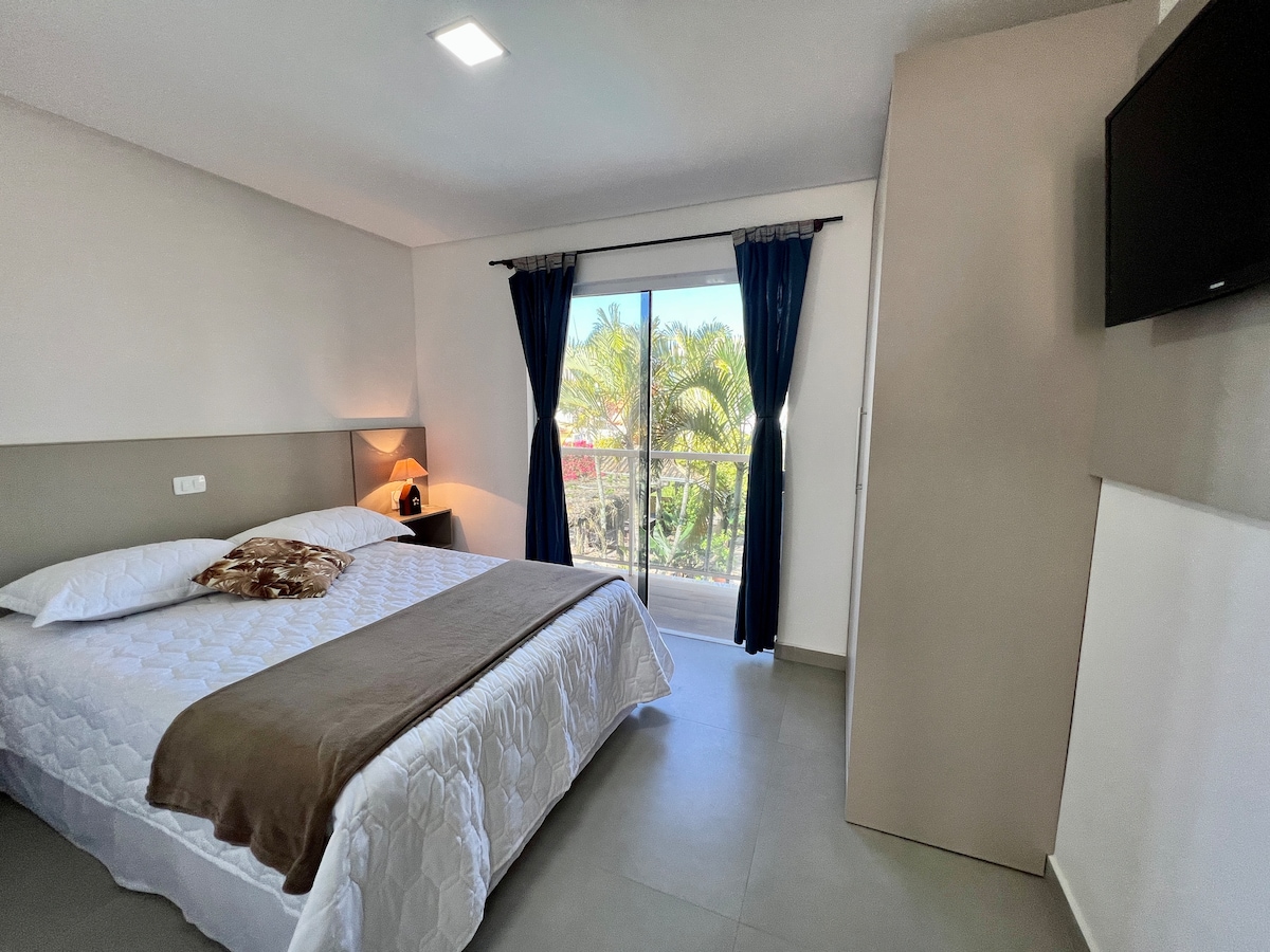 A bright and airy bedroom is featured, showcasing a spacious bed draped in white linens with a brown throw. Large windows framed by dark curtains allow natural light to filter in, providing a view of greenery outside. A television is mounted on the wall.