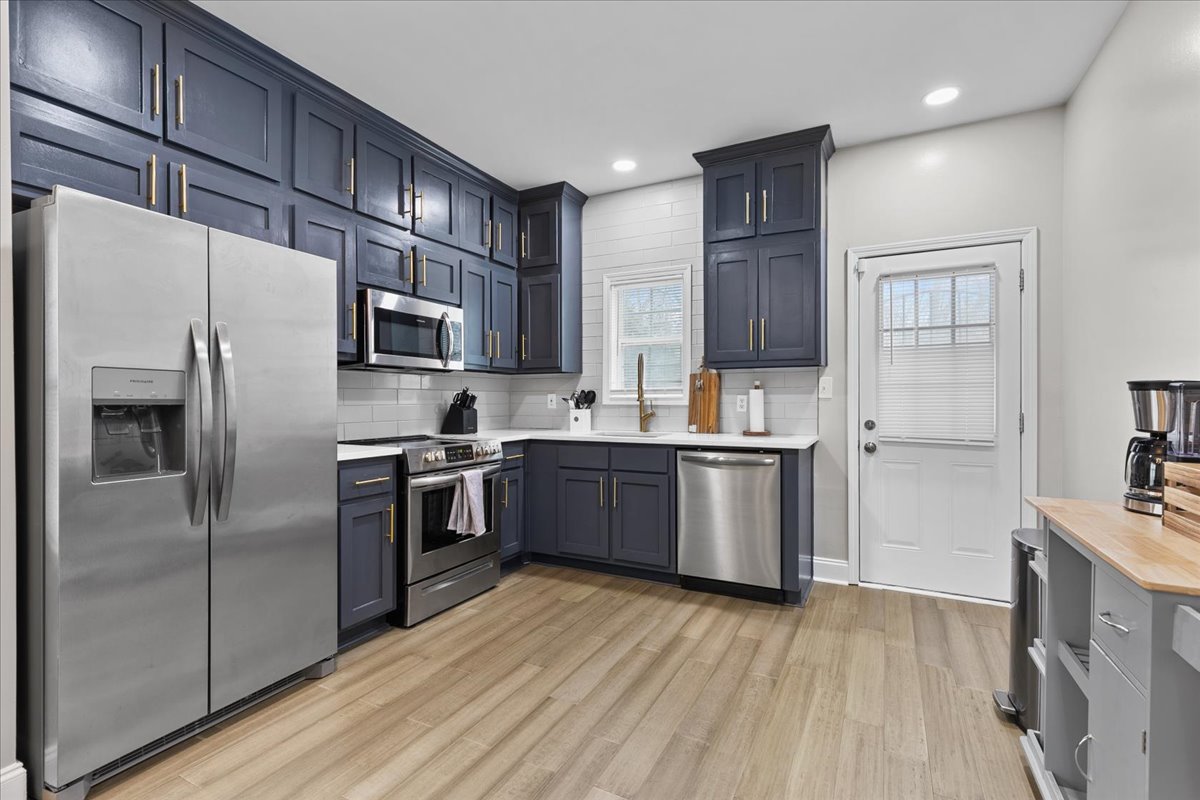 A modern kitchen is equipped with stainless steel appliances, including a large refrigerator and a range oven. Dark cabinetry contrasts with the light countertops, while ample natural light enters through a nearby window. A door leads to the exterior, enhancing accessibility.
