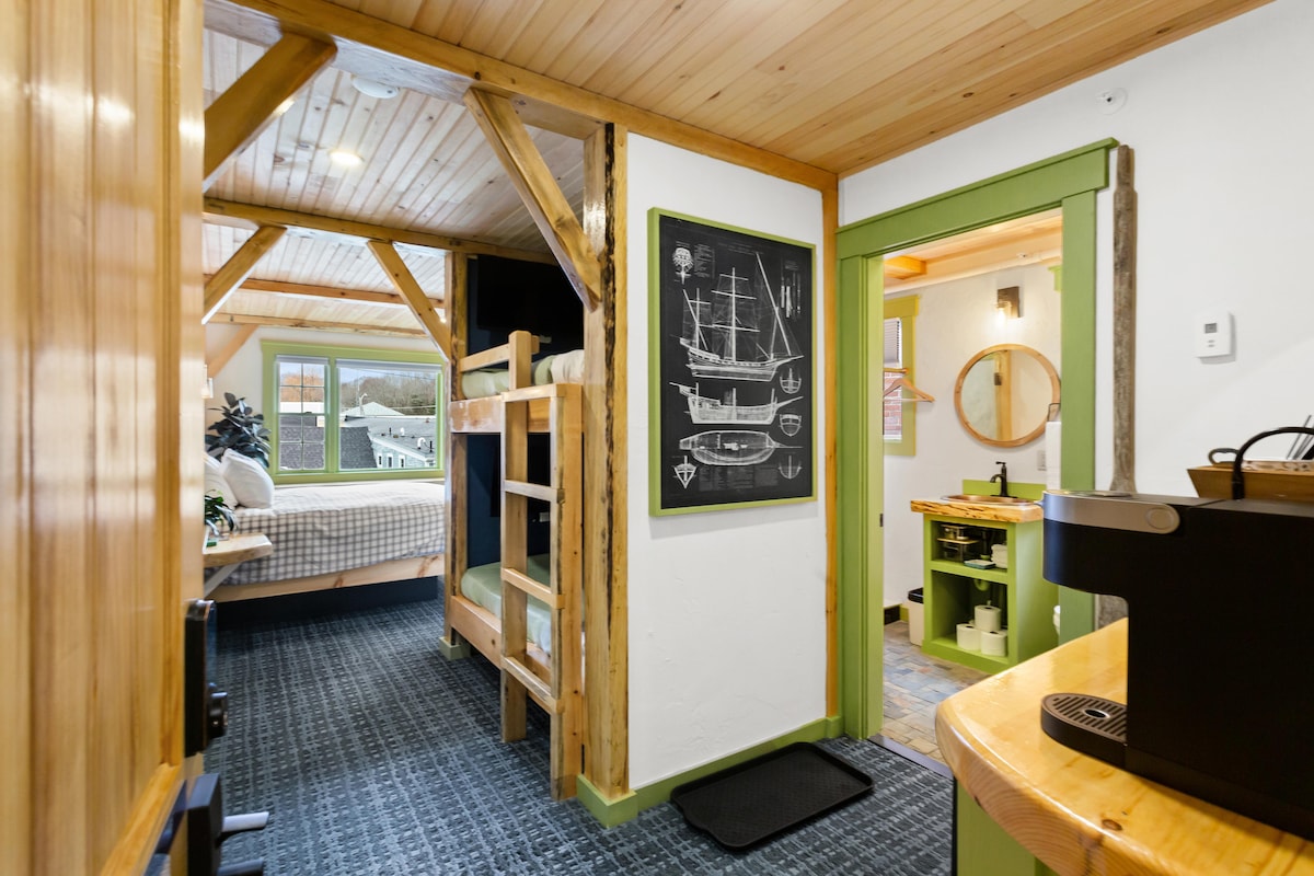 The room features wooden beams and a warm, welcoming atmosphere. A custom bunk bed designed for children is visible alongside a cozy king-sized bed. The kitchenette is equipped with modern amenities, and a round mirror enhances the space. Natural light spills in from spacious windows.