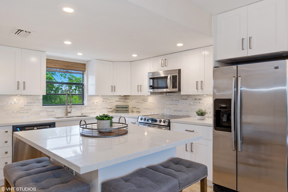 A modern kitchen features white cabinetry and stainless steel appliances, including a refrigerator and microwave. A spacious countertop is complemented by a circular tray with decorative items. Soft natural light enters through a window, enhancing the clean and organized space.
