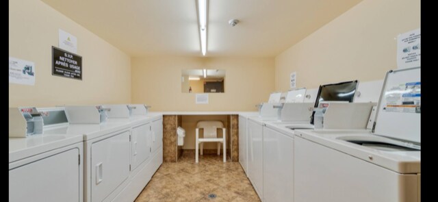 A laundry room is presented, featuring multiple washing machines and dryers arranged in a row. A folding table is situated centrally, accompanied by a white chair. The walls are painted in a neutral tone, and overhead lighting provides ample illumination.