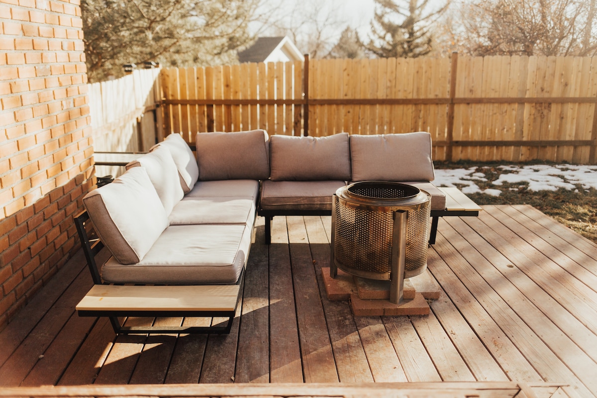 A spacious outdoor seating area is presented on a wooden deck, featuring a sectional sofa with soft cushions. A round fire pit sits at the center, surrounded by wooden planks. A fenced yard with grass and scattered snow is visible in the background.