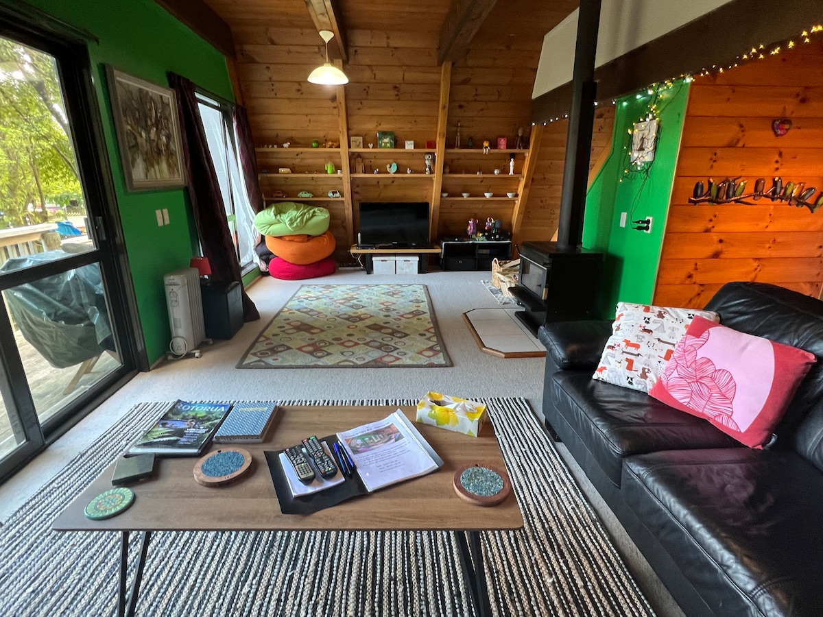 A vibrant living area features wooden beams and green walls, with a cozy black sofa and a coffee table adorned with magazines and stationery. A bright rug adds warmth, while a shelf displays decorative items. Large glass doors reveal access to an outdoor deck.