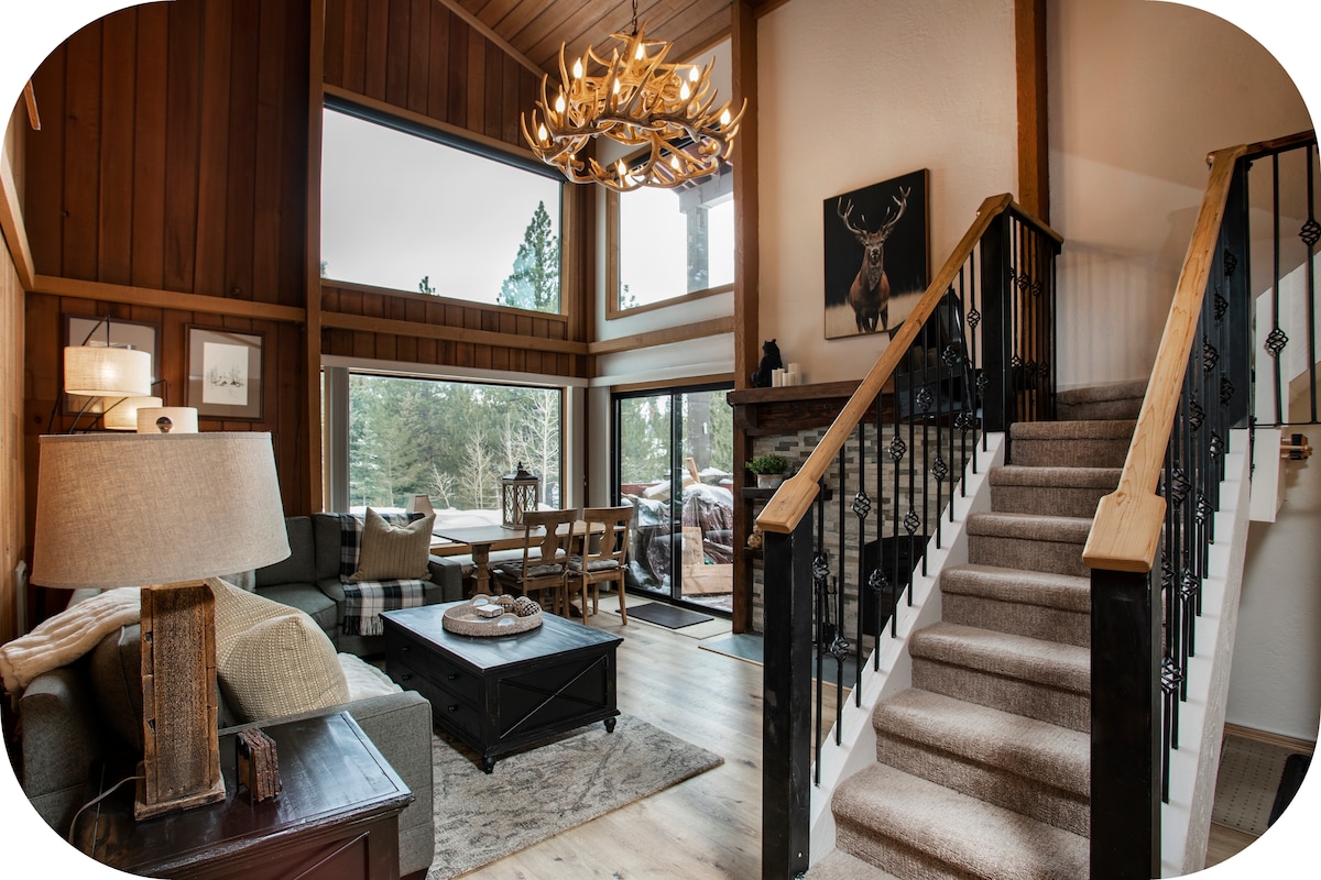 A warm living area features wood-paneled walls and large windows offering natural light. A chandelier hangs from the ceiling above stylish furnishings, including a comfortable sofa and a coffee table. Stairs lead to an upper level, while a cozy fireplace is positioned against a backdrop of rich woods.