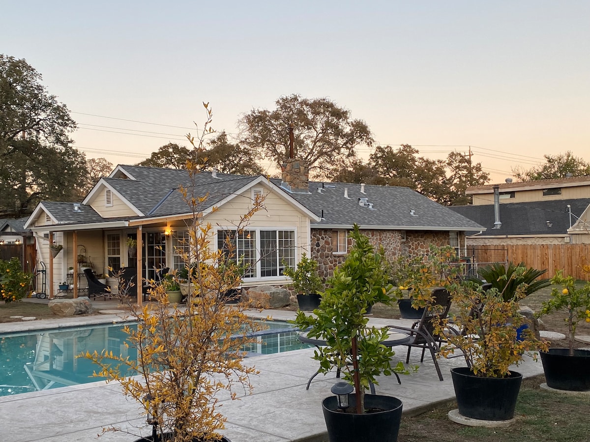 A spacious backyard features a large pool surrounded by well-maintained landscaping. Potted plants and fruit trees are strategically placed around the area. The home is partially visible in the background, showcasing a warm glow from the evening light.