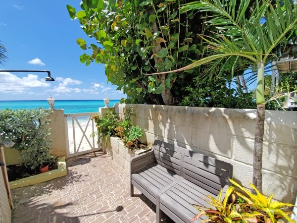A private outdoor seating area features grey benches surrounded by lush green foliage. A stone pathway leads to a gated entrance, with the sandy beach and turquoise waters visible just beyond a low wall. Bright sunlight illuminates the scene.