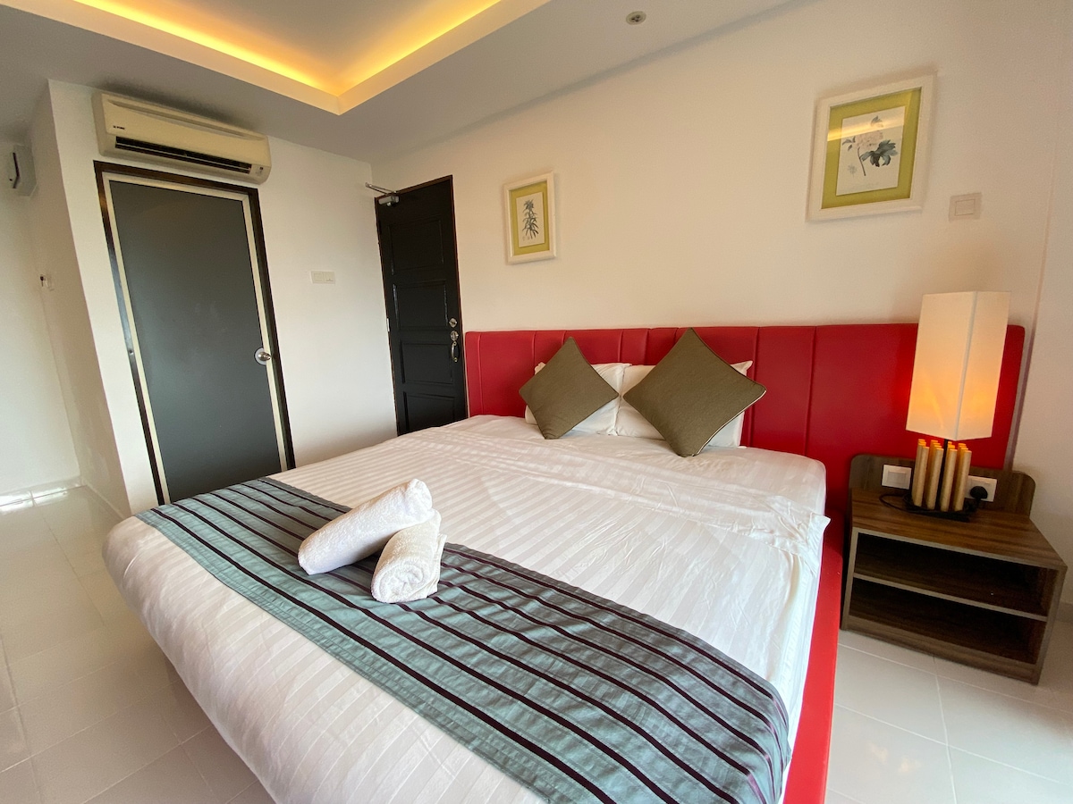 A spacious bed is positioned against a vibrant red headboard, complemented by decorative pillows. A wooden nightstand with a lamp is nearby, and artwork is displayed on the white walls. Soft lighting creates a calming environment, while a door leads to the outside.