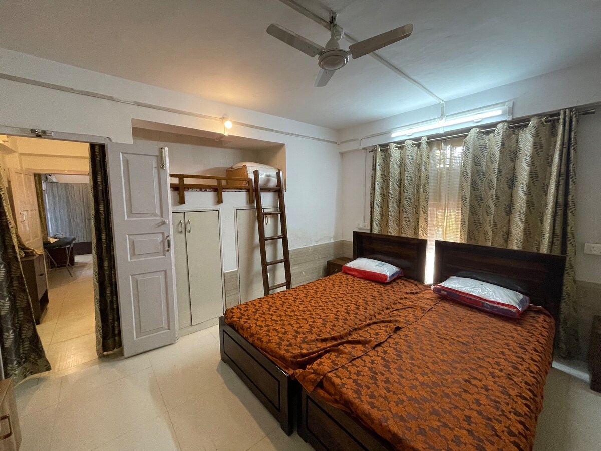 The interior space features a well-lit room with two spacious beds covered in orange bedding. A wooden ladder leads to an upper sleeping area, and a wardrobe is positioned against the wall. Light filters through partially drawn curtains.