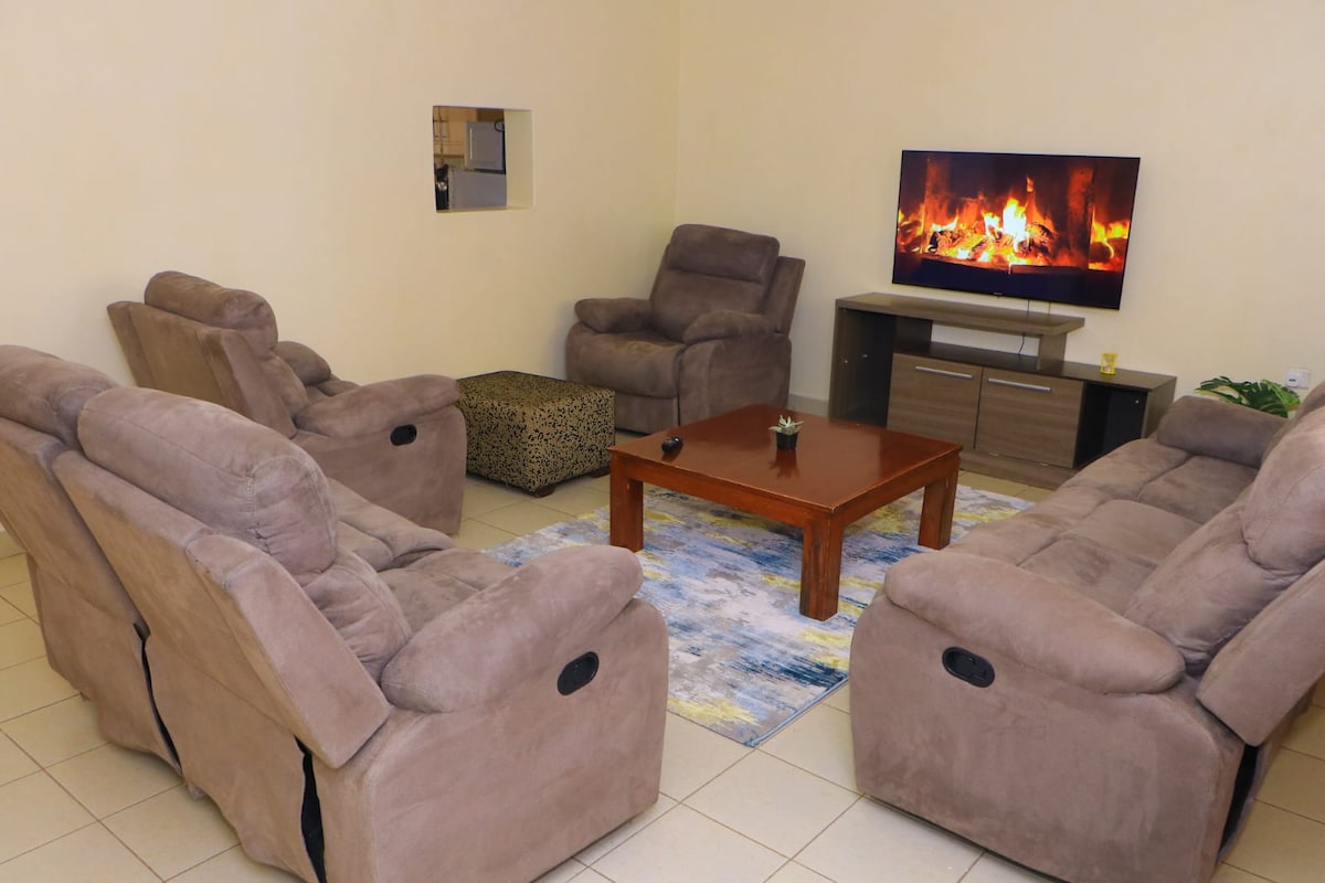 A relaxed living area features four reclining chairs arranged around a wooden coffee table. A television mounted on the wall displays a fireplace scene, contributing to a cozy ambiance. A patterned rug adds visual interest underfoot.