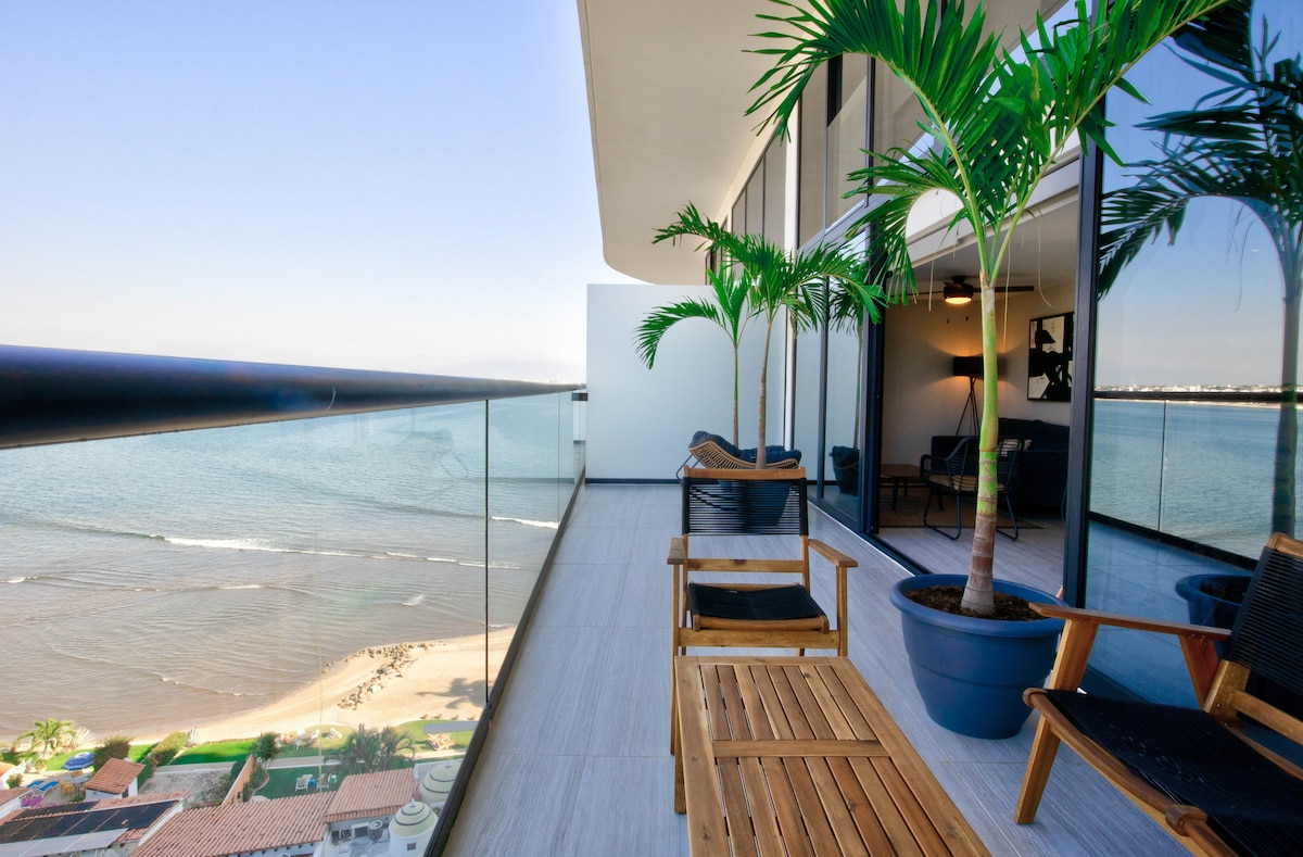 A spacious balcony offers sweeping views of the ocean, complemented by two wooden lounge chairs and potted palm trees. The glass railing provides an unobstructed perspective of the sandy beach below, creating a serene setting for relaxation.