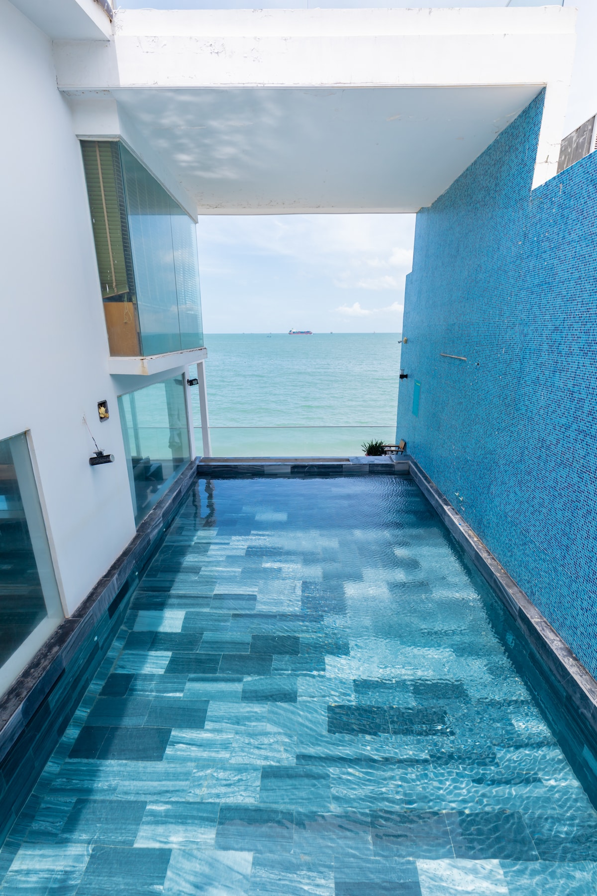 A clear swimming pool is visible, reflecting the sky and ocean, with varying shades of blue tiles creating a serene design. The pool overlooks the water and is framed by a modern building featuring large windows and a vibrant blue wall.