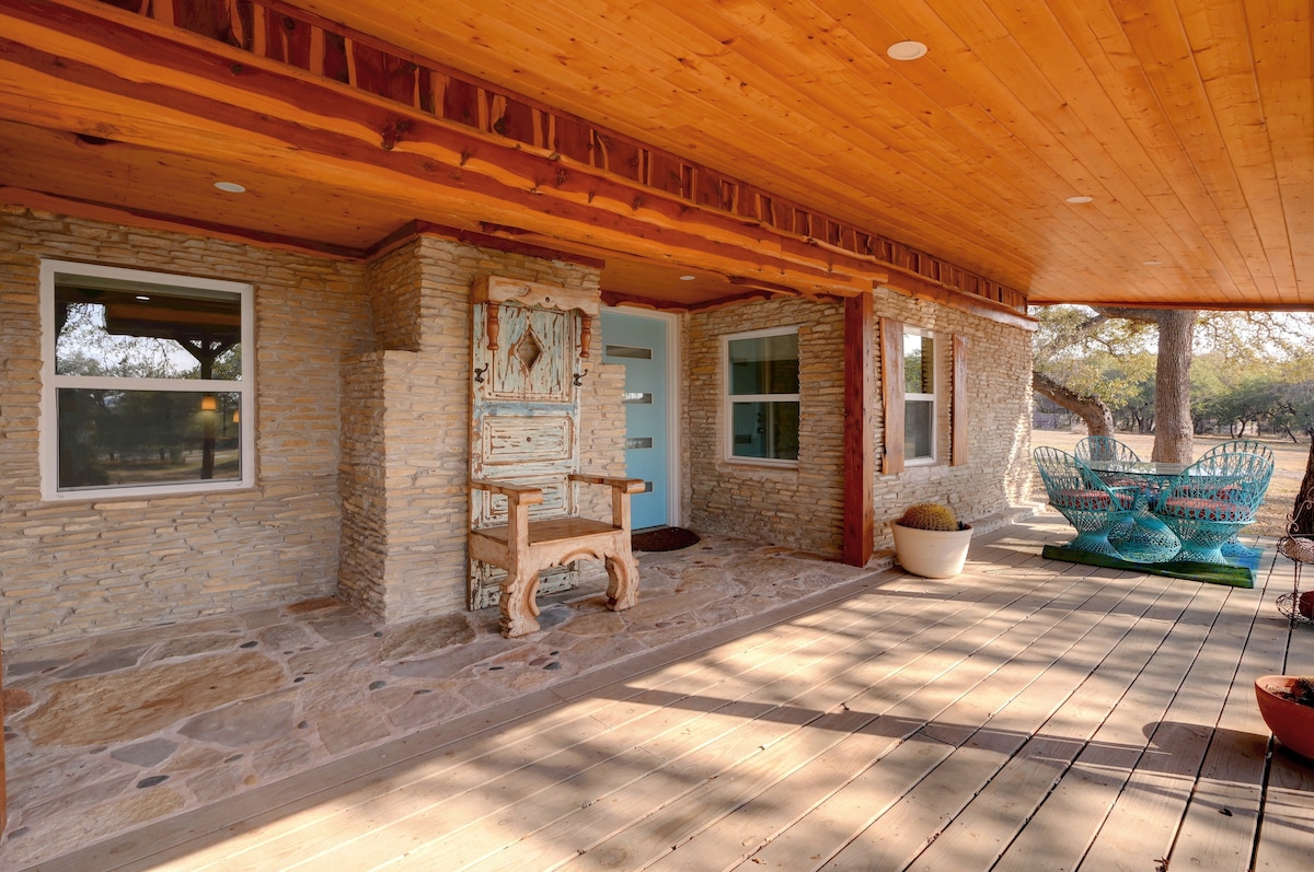 A front porch features natural stone walls and wooden ceilings, creating a welcoming entrance. Seating in a unique turquoise design is complemented by potted plants. Ample natural light filters in through the windows, illuminating the space decorated with rustic elements.