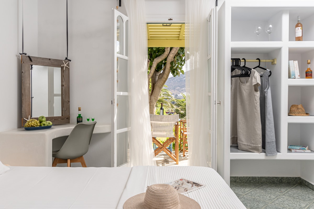 A bright studio is featured, with light entering through open French doors that lead to a private terrace surrounded by greenery. A small dining area with a modern chair is visible, alongside shelves displaying clothing and personal items, creating a functional and inviting space.
