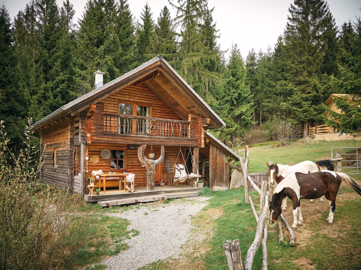 A rustic wooden cabin features a spacious porch with a dining area, flanked by decorative elements. Two horses graze nearby on a grassy area, with tall trees providing a natural backdrop. The structure showcases a warm, inviting aesthetic in harmony with its natural surroundings.
