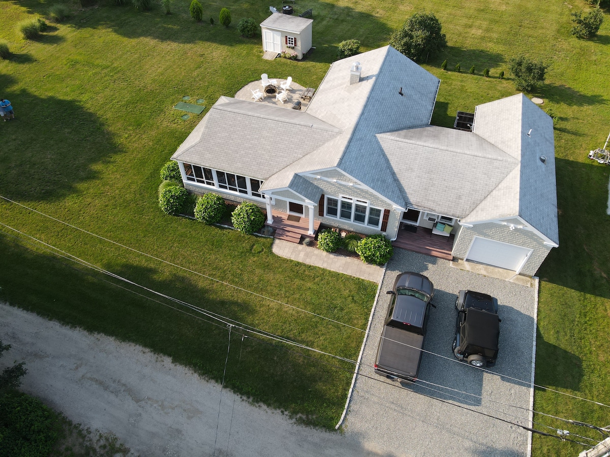 An aerial view of a spacious single-story home is displayed, featuring a large front yard and a gravel driveway accommodating multiple vehicles. The house has a screened-in porch and a deck entryway, with greenery outlining the property, enhancing the outdoor setting.