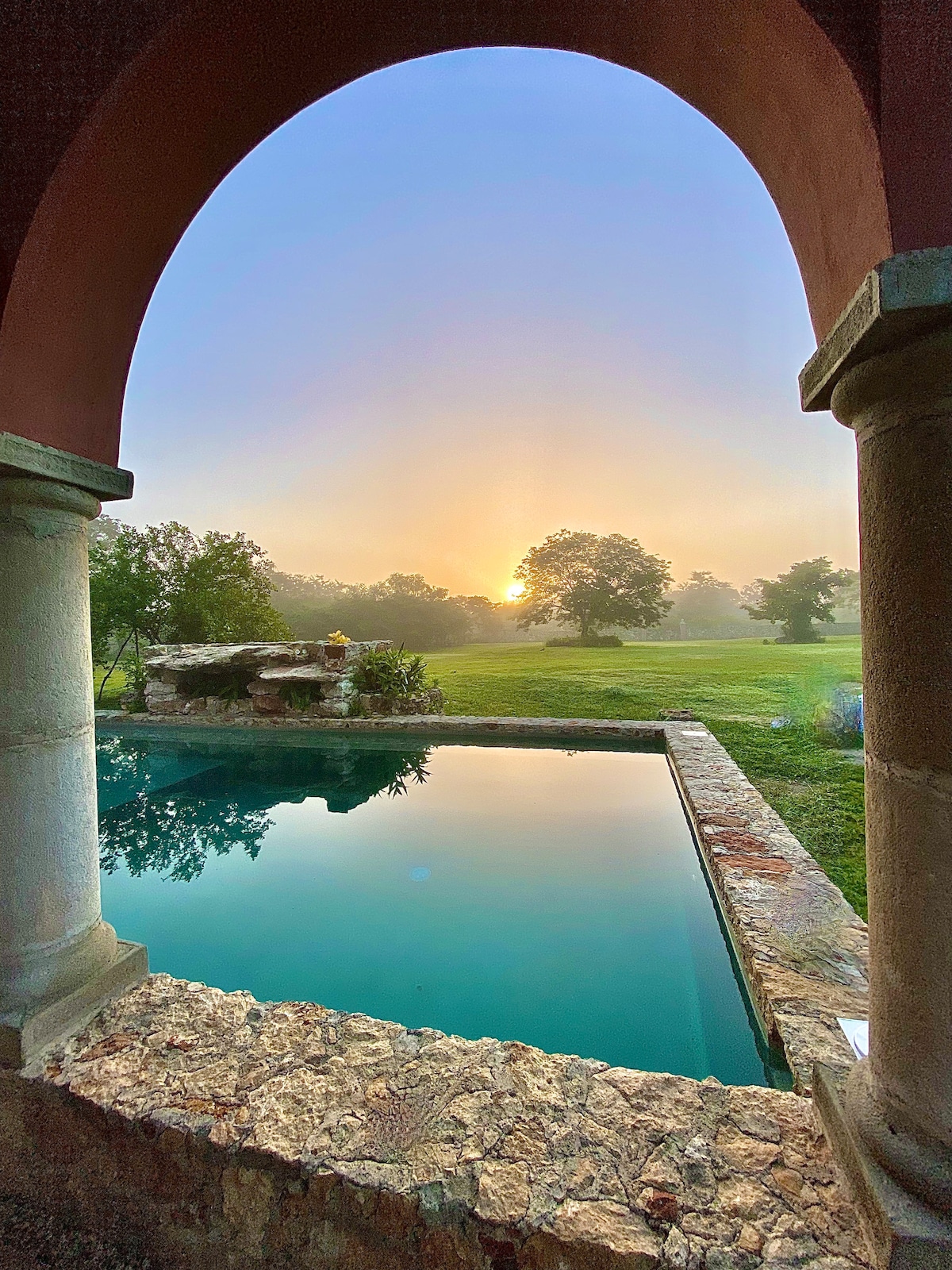 A tranquil pool reflects the soft morning light, framed by an archway and surrounded by lush greenery. The rising sun casts a gentle glow over the serene landscape, creating a peaceful ambiance in this secluded outdoor space.