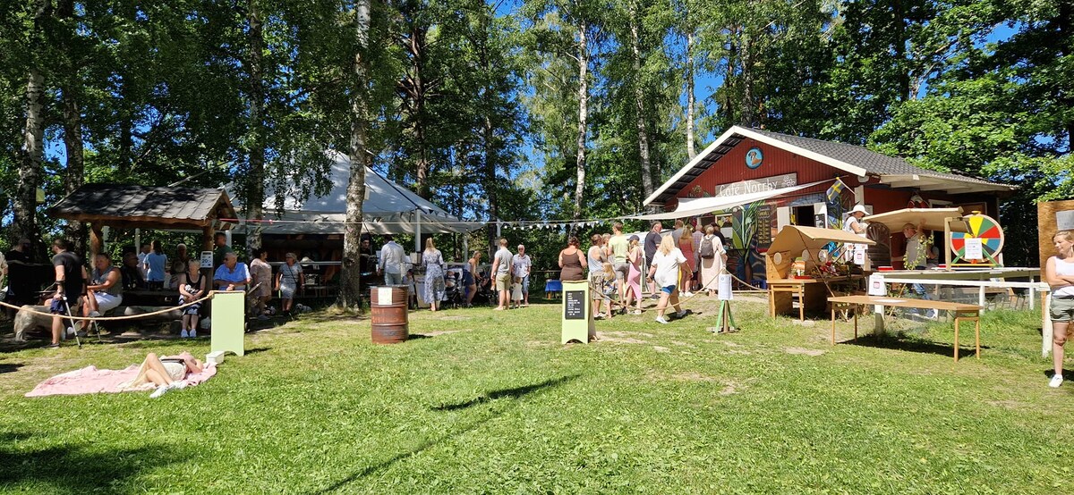 A vibrant outdoor gathering is depicted, featuring a spacious lawn surrounded by tall trees. A red building is visible in the background, along with a white tent. Guests enjoy various activities, while a picnic setup and wooden barrels are present on the grass.