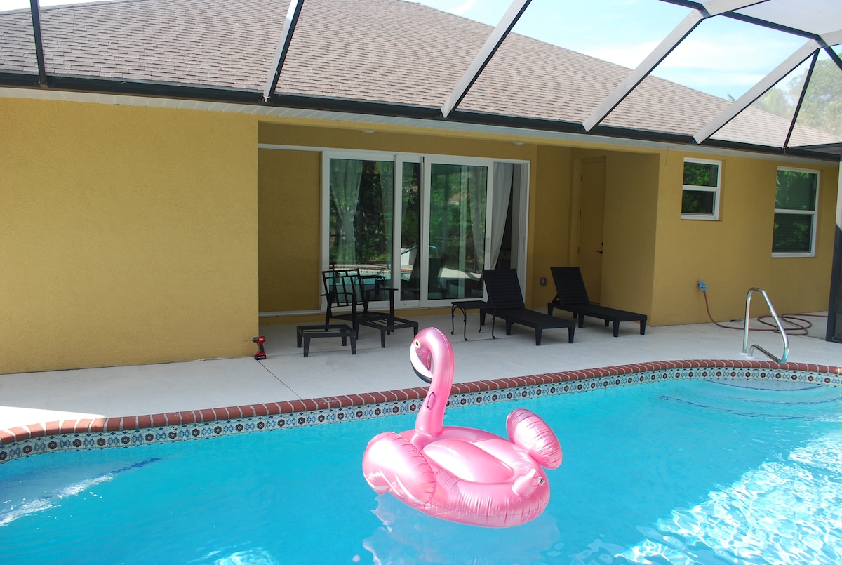 A private pool area is featured, complete with a vibrant pink flamingo float. Lounge chairs are positioned along the edge of the pool. Large glass doors provide a view of the indoor space, while sunlit concrete surrounds the water.