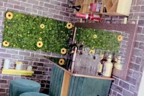 A stylish bar area is highlighted by a backdrop of artificial greenery adorned with sunflower accents. The countertop is lined with various bottles, and colorful bar stools are present. Bricks in a warm tone add to the contemporary aesthetic of the space.