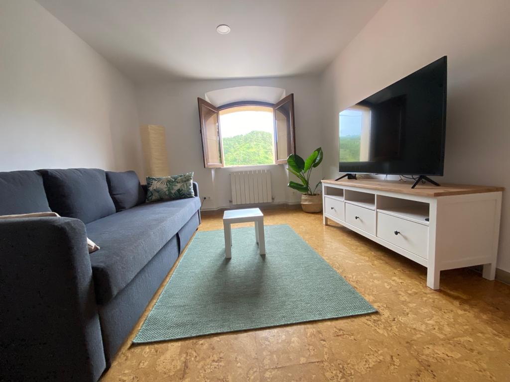 A living area features a dark gray sofa positioned near a window that provides mountain views. A small white table rests on a green rug, with a light-colored TV cabinet displaying a flat-screen television. Natural light brightens the space, enhancing its airy feel.