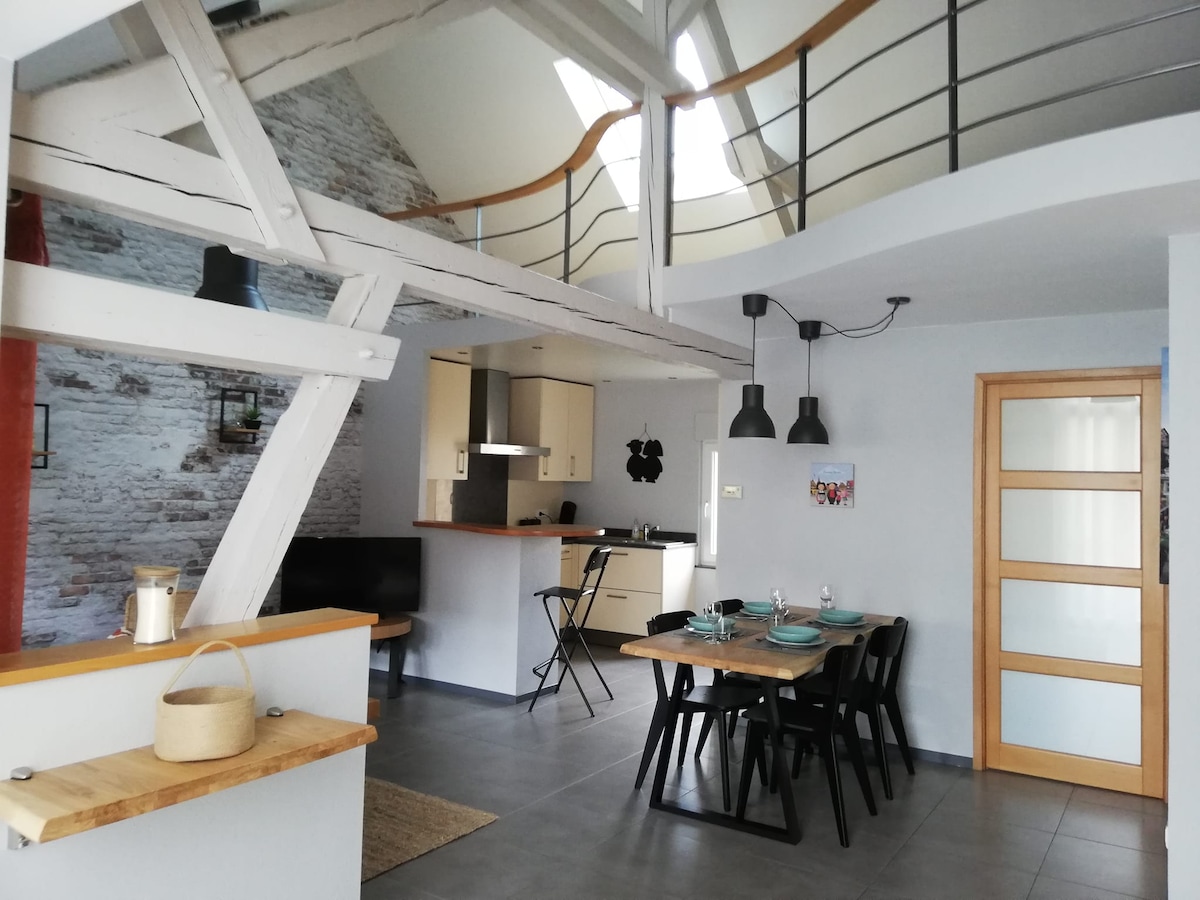 The open living area showcases exposed beams and high ceilings, with a dining table set for six. The kitchen features modern appliances and a breakfast bar. A cozy seating area with a dark sofa invites relaxation, while natural light filters through the large windows.