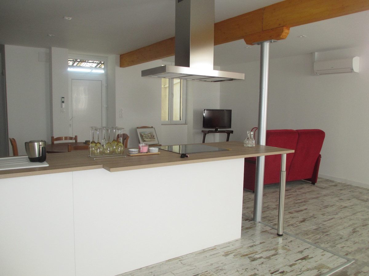 An open kitchen is featured with a central island and modern appliances. A dining area with wooden chairs is visible. A cozy living space includes a red sofa and a small television. Bright light enters through large windows, enhancing the airy ambiance.