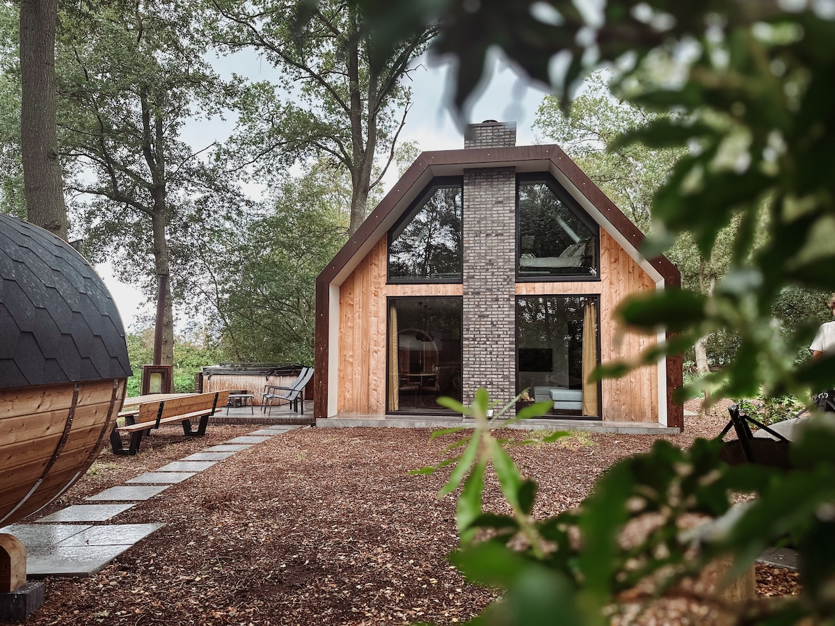 The modern house, constructed from CorTen steel, showcases large glass windows framed by wooden siding. A spacious terrace features outdoor seating, and a pathway leads to the entrance. Natural surroundings include tall trees providing shade, enhancing the serene atmosphere.