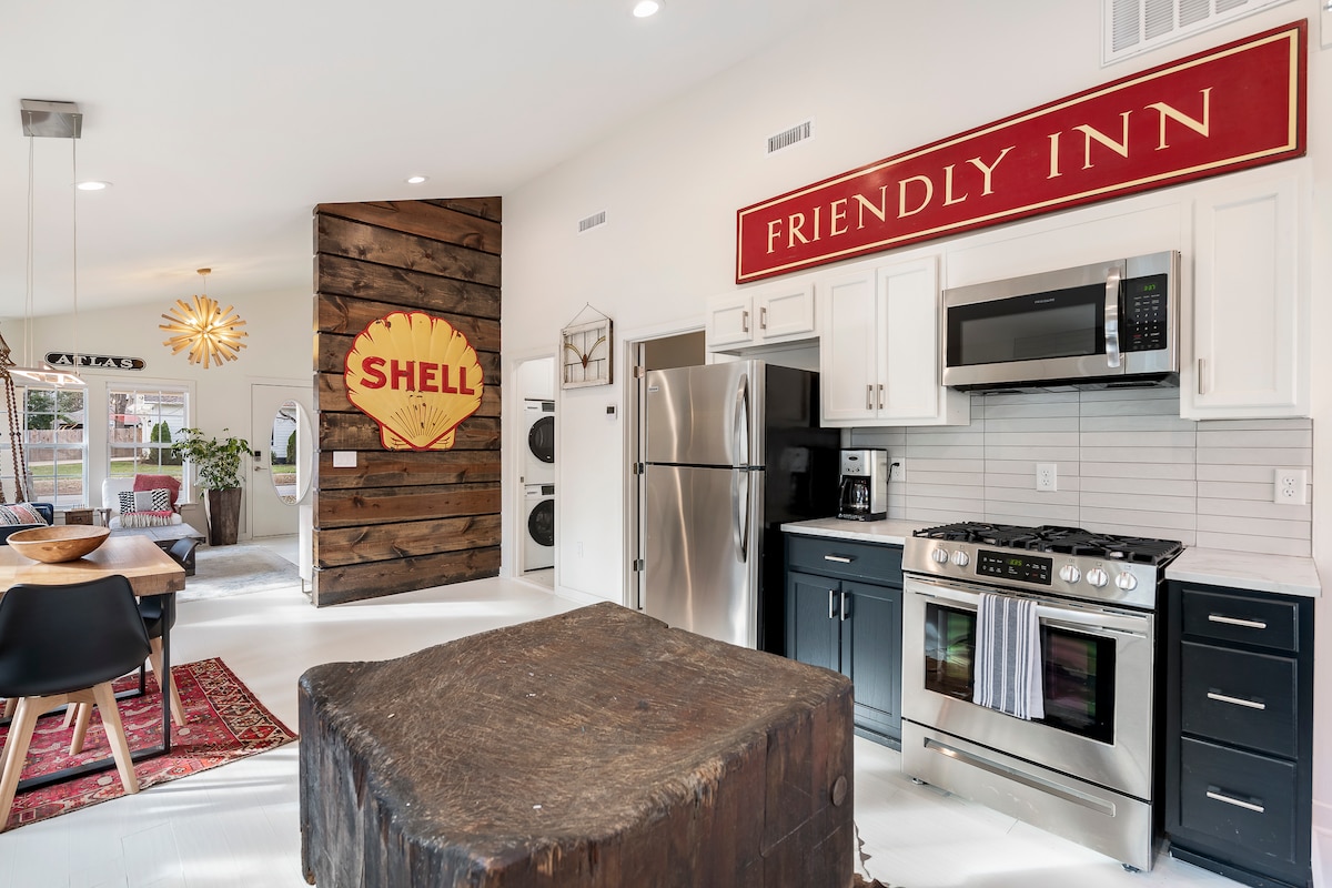 An open kitchen area is highlighted by a large, rustic island made of dark wood. Modern appliances, including a stainless steel refrigerator and oven, are present. A vibrant sign reading 'FRIENDLY INN' adds character to the wall, while a dining area with a round table is visible in the background.