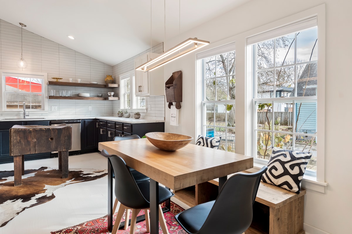 A modern kitchen area features a wooden dining table complemented by sleek black chairs. Natural light enters through multiple windows, illuminating the space. A stylish light fixture hangs above the table, and open shelving displays kitchenware, while a decorative bowl rests at the table's center.